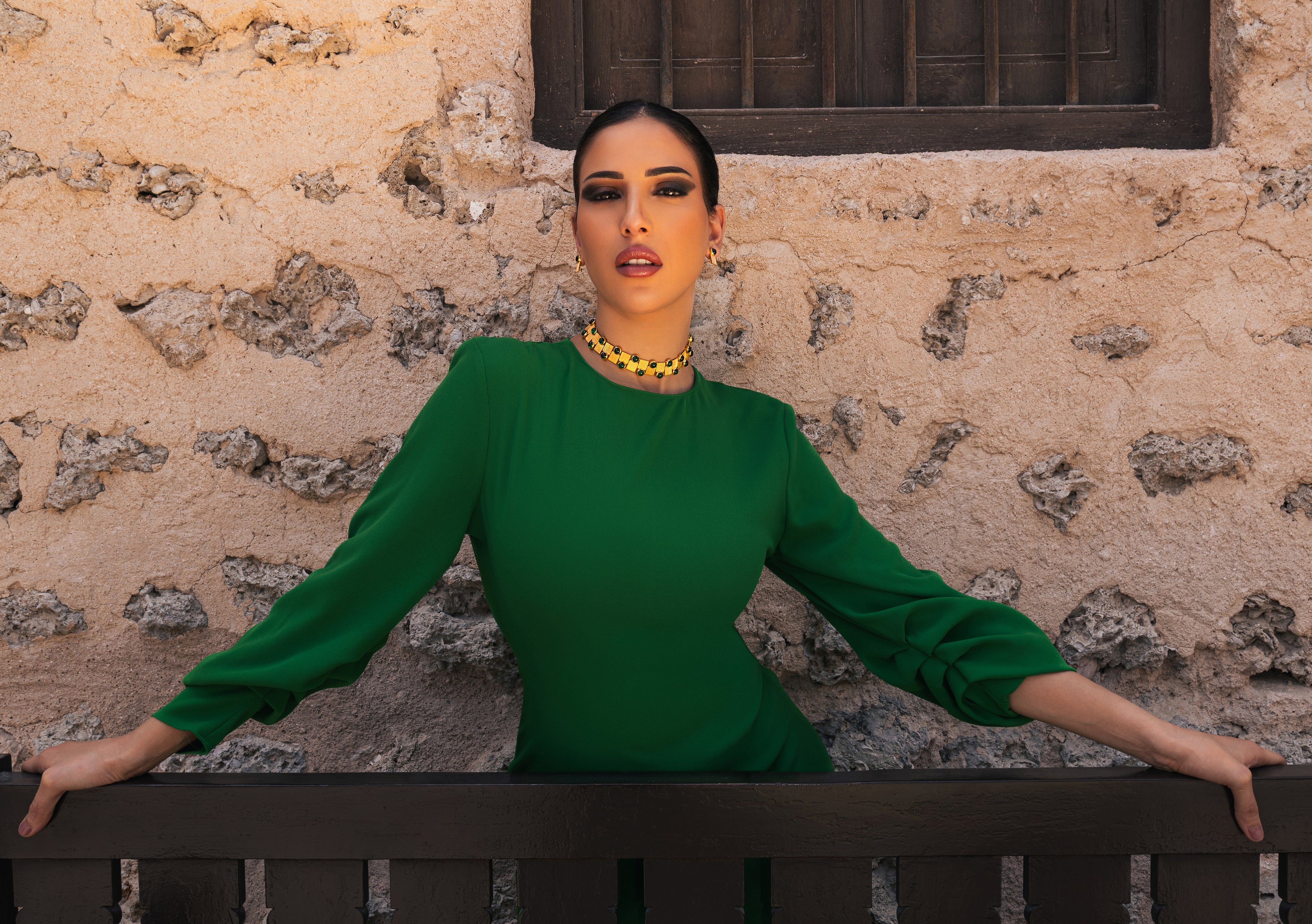 Woman in a green dress standing against a stone wall with jewelry